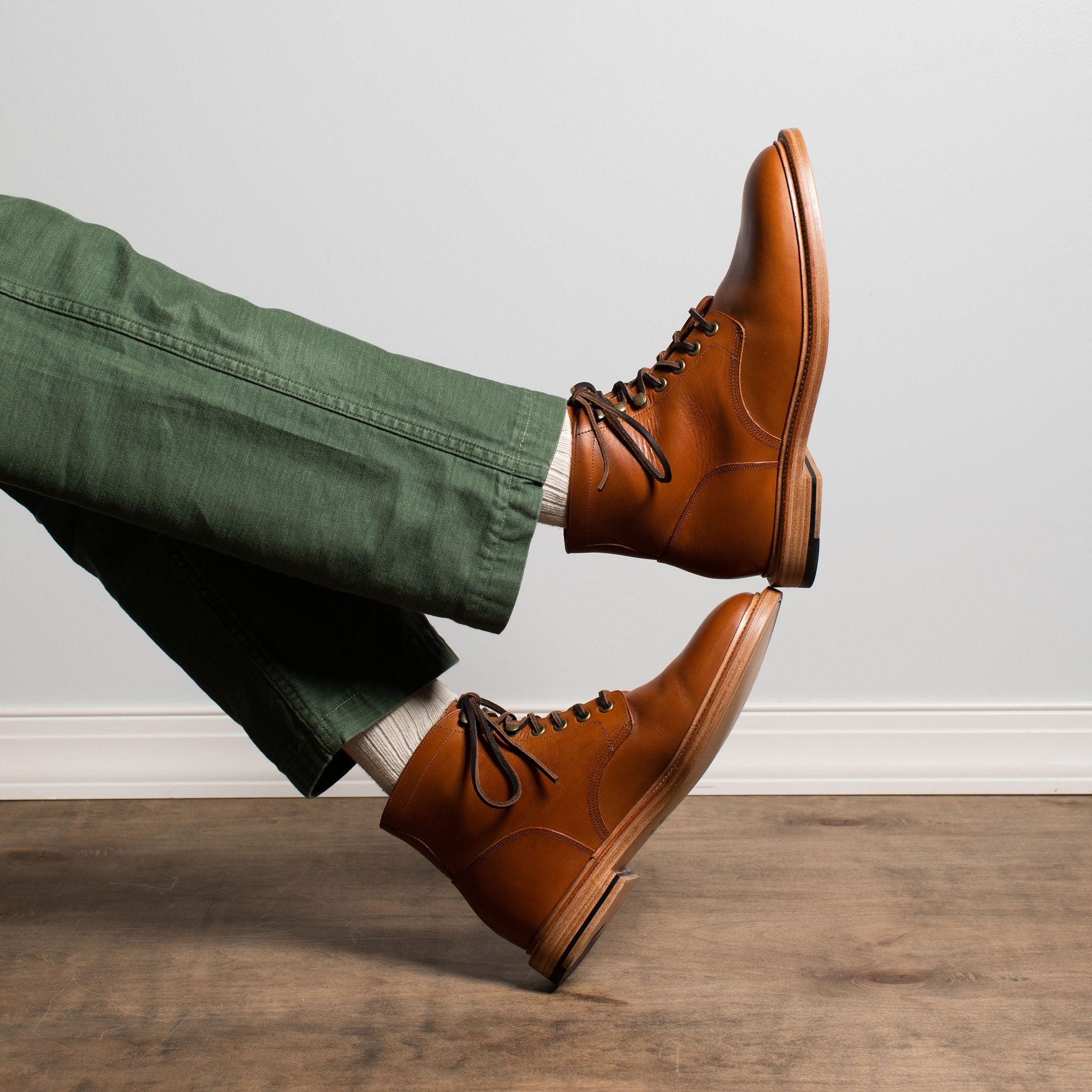 A person wearing green pants and white socks with a pair of brown leather lace-up boots, posed with legs crossed on a wooden floor against a light gray wall.