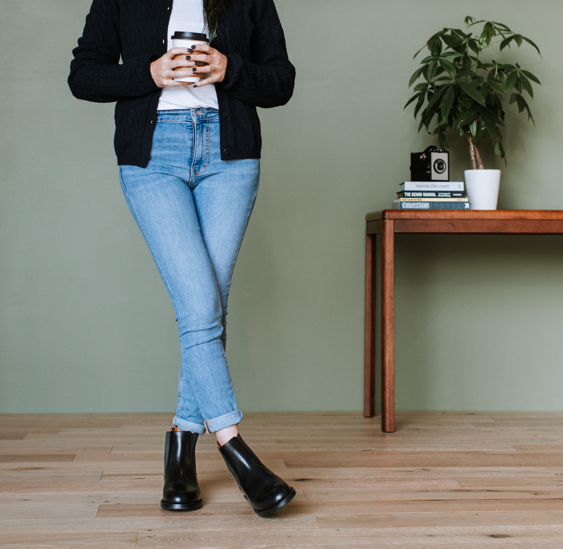 A person wearing light blue jeans, a black cardigan, a white top, and black ankle boots stands indoors against a muted green wall, holding a takeaway coffee cup. A wooden table with books and a potted plant is visible on the right.