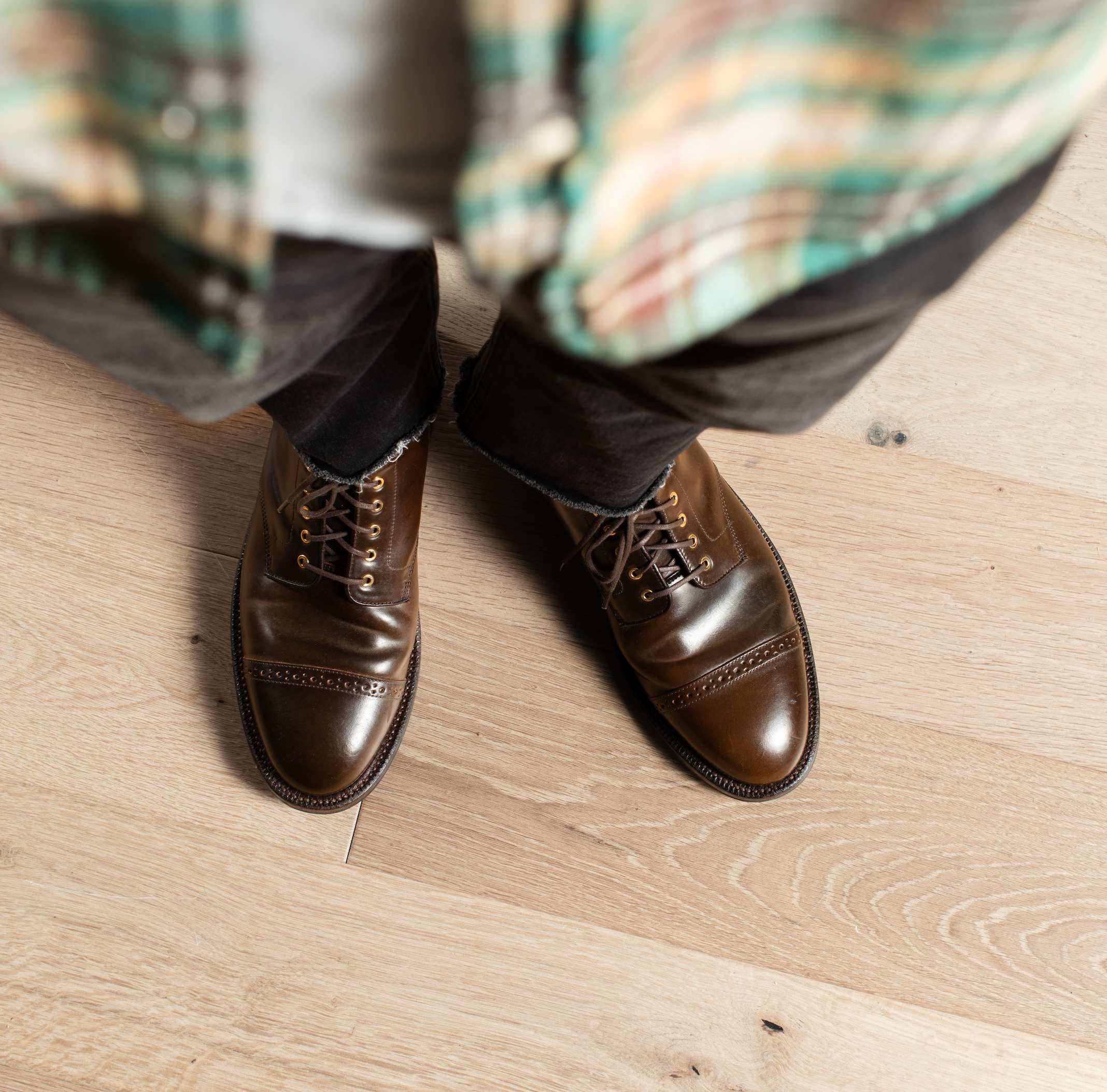 A close-up, high-angle shot of a person wearing a pair of polished brown leather wingtip cap-toe oxford shoes with brogue detailing and laces, standing on a light-colored wood plank floor. The person's legs are visible, wearing dark trousers and a green and yellow plaid shirt.